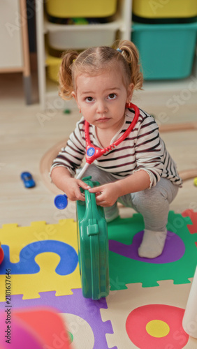 Foto Little blonde girl squats holding a green toy suitcase while wearing a red toy stethoscope and playing on foam puzzle mats in a bright playroom; curiosity