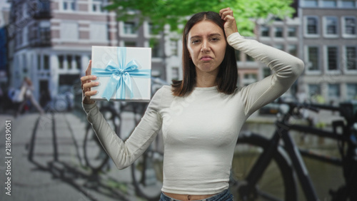 Young woman holds gift box with blue ribbon in left hand and scratches head with right, grimacing on a city street; confusion hesitation.