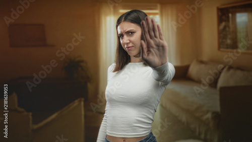 Fotografie Young woman shows palm raised in a stop gesture inside a building living room; defiance boundary refusal strength