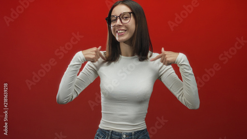 Young woman points both fingers to chest and smiles, wearing glasses in a red studio; confidence positivity.