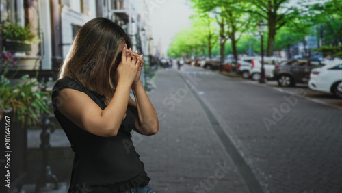 фотографія Woman covers mouth with hands and wears glasses while standing on a tree lined street near parked cars; surprise privacy vulnerability