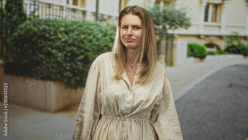 Foto Blonde woman standing on street in beige linen dress grimacing with pursed lips showing face and necklace; skepticism awkwardness