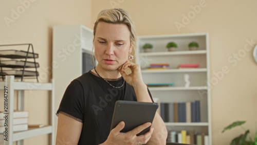 Bild auf Leinwand Woman holding tablet, hand to mouth while reading in an office building with bookshelves and a desk, looking down at screen; concerned reflection
