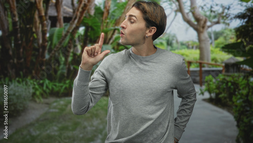 Foto Man in grey sweater holds index finger up while looking sideways on a leafy forest path beside a wooden railing; thoughtful warning