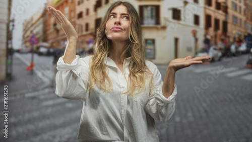 Bild auf Leinwand Woman in white shirt raises both palms in a shrug gesture and looks upward on a cobblestone street; puzzlement reflection