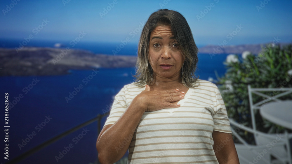 Obraz premium Woman middleaged hispanic with hand on chest looking up on building balcony with white railing, striped shirt and sea view; quiet gratitude.