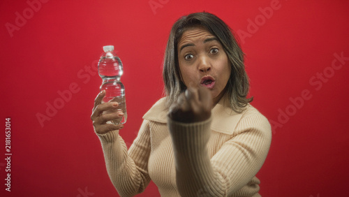 фотографія Middle age hispanic woman holding water bottle and pointing fist to camera in red studio, leaning forward with expressive face; assertive confidence