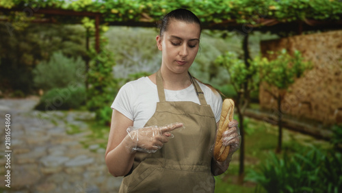 Canvastavla Woman in apron holding a baguette and pointing a finger with gloved hand on a stone path in building courtyard; annoyance food safety