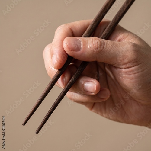 Close up of a hand holding traditional dark wooden chopsticks against a minimalist neutral background for Asian dining presentation