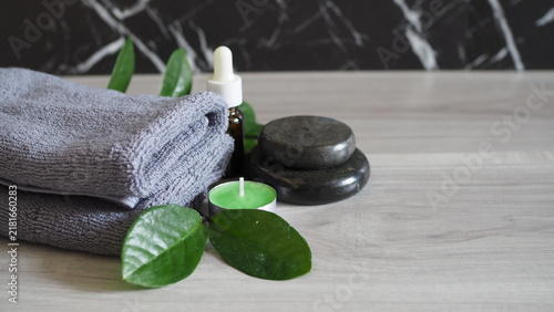 Canvas Print Spa wellness arrangement featuring folded gray towels, smooth dark massage stones, a serum dropper bottle, an unlit green tealight candle, and fresh green leaves on a light wooden background