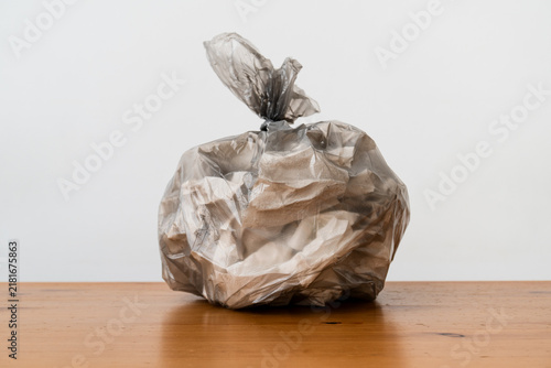 Close-up photo of tied transparent plastic bag filled with household trash on wooden table. Crumpled packaging and waste are visible through bag. Image conveys garbage disposal and consumption.