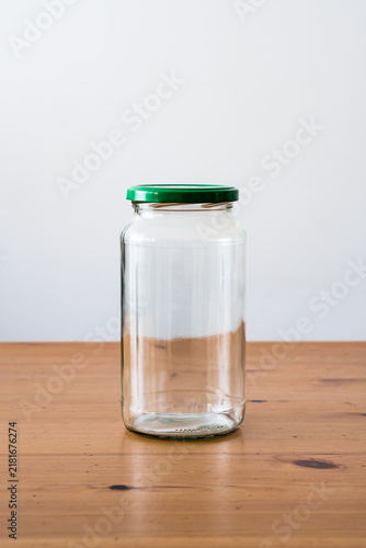 Front view of empty clear glass jar with green metal lid on wooden table against minimalist light grey background with copy space.