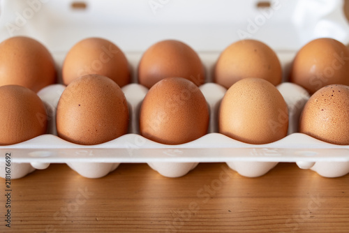 Close-up photo of open egg carton holding ten brown eggs on wooden kitchen table. Authentic image of pack of ten eggs fresh out of the fridge made in the kitchen.