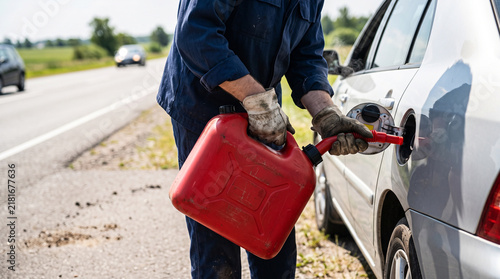 Man in blue overalls pouring fuel from a red Jerry can into a car on a highway shoulder.