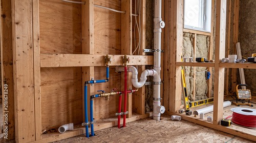 Plumbing installation in a residential construction site showing red and blue PEX water lines, PVC drainage pipes, and wooden wall framing.