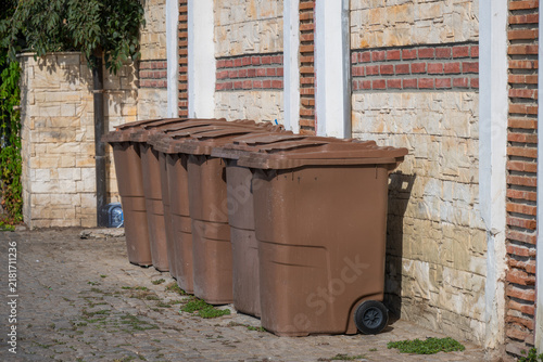 Konstfotografi A row of identical brown plastic garbage bins stands lined up on a cobblestone surface against a light stone and brick wall