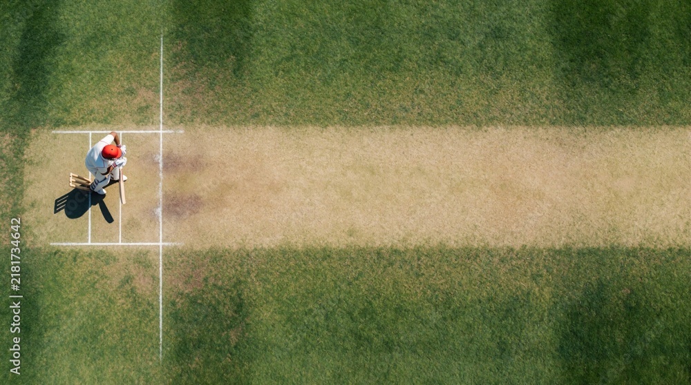 Obraz premium Top view of cricket pitch on green grass field. Male athlete wears white uniform, red helmet preparing to bat. Player holds wooden bat at crease during sunny daytime match in sports stadium for