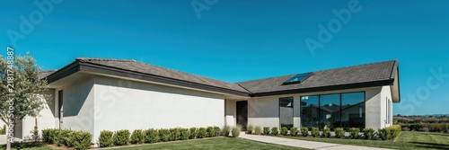 Contemporary L-shaped house with stucco finish large windows and modern roof against bright blue sky for luxury living.