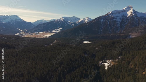 Aerial view of snow-capped peaks above dense forests. Sunlight glints off distant mountain summits