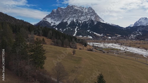 Golden fields stretch toward snow-capped peaks under a bright sky. Firm, leafless trees dot the landscape in quiet harmony