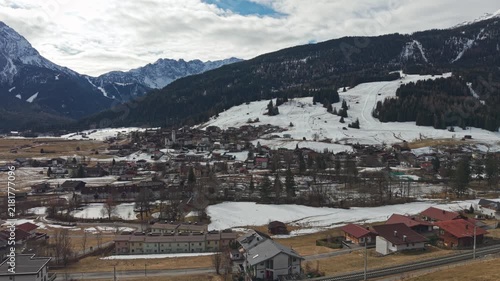 Snow-dusted mountains frame a quiet village below. Roads wind through homes with red-tiled roofs