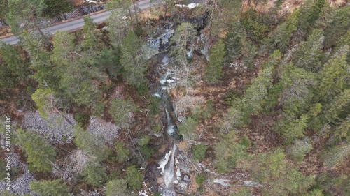 Aerial view of a narrow stream rushing through forested terrain. Road runs parallel, bordered by dense evergreen trees