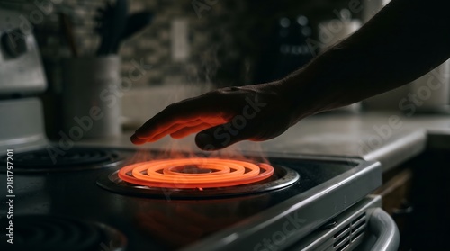 A close-up of a hand hovering over a glowing red electric stove burner, testing the intense heat in a dimly lit kitchen.