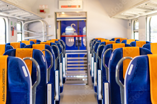 Empty train carriage interior shows rows of blue and orange seats stretching toward the front. Concept of modern public transportation and clean train interior design for travel content.