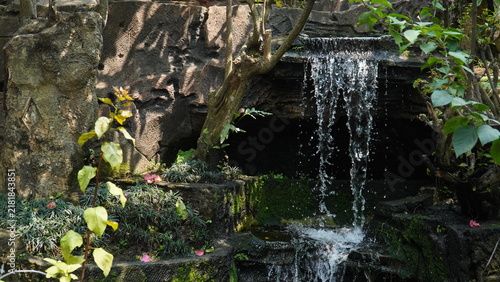 Photography A small decorative waterfall flows over dark rocks in a lush green garden setting during daytime