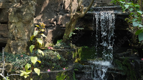 A small decorative waterfall flows gracefully over tiered rocks in a lush tropical garden setting.