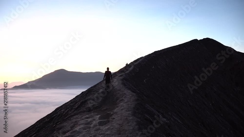 Man in silhouette hiking along a volcanic crater rim at sunrise with a sea of clouds below