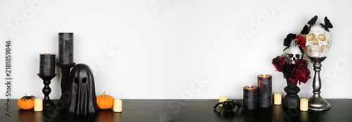 Halloween gothic shelf decor with copy space against a white wall banner background. Black shelf with glowing candles, dark flowers, skulls, pumpkins, ghost and spiders.