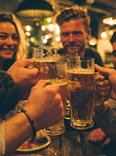 Radosna grupa przyjaciół uśmiecha się, wznosząc toast kufelkami pełnymi złocistego piwa w ciepłej, nastrojowej scenerii wieczornego spotkania.