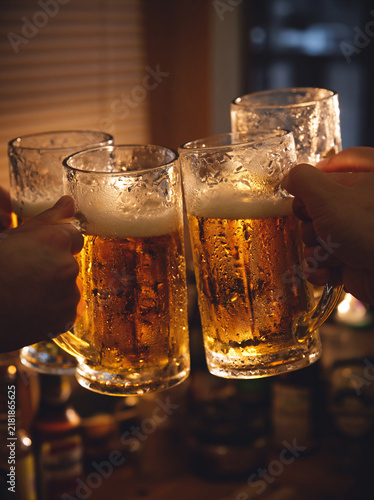 Grupa przyjaciół wznosi toast, trzymając w dłoniach ciężkie szklane kufle wypełnione schłodzonym, złocistym piwem z gęstą pianą. W tle widać nastrojowe, wieczorne światło klimatycznego wnętrza.