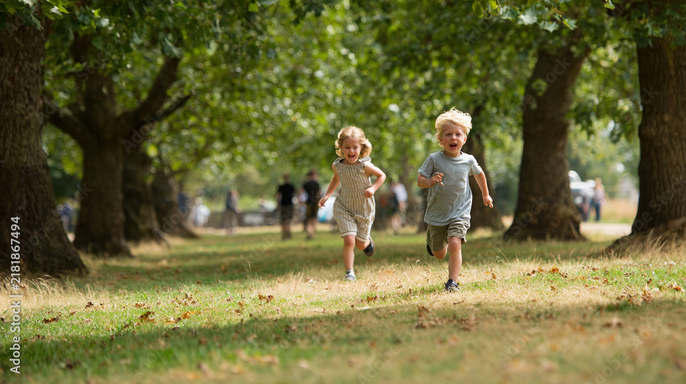 Fototapeta premium Happy young children running together through green park trees during a bright warm summer day outdoors
