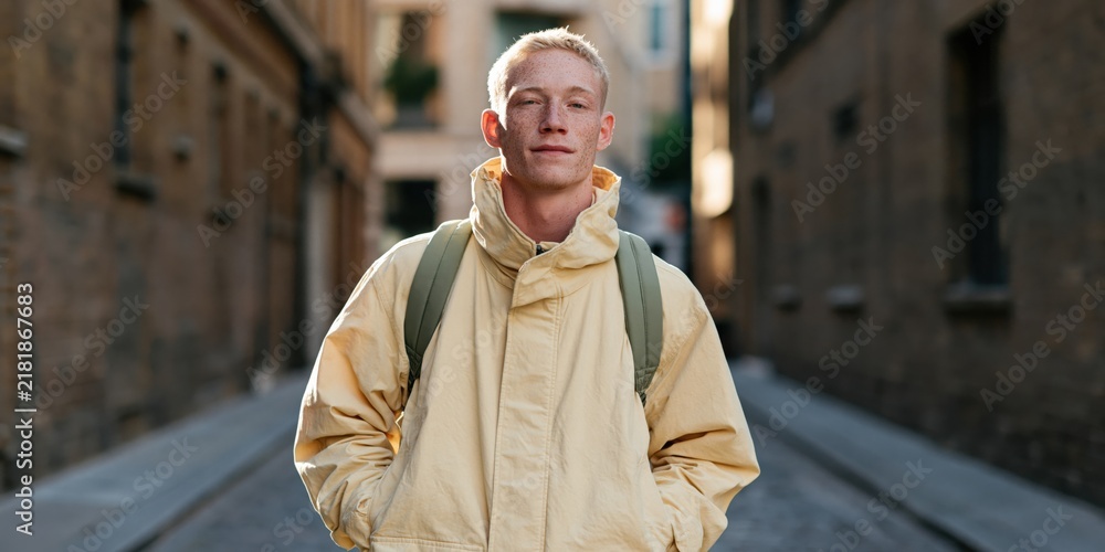 Obraz premium Young man in pale beige jacket with backpack standing in a narrow urban alley for portrait background and banner use