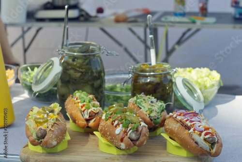 Close-up of gourmet hot dogs with fresh toppings on a rustic wooden board, pickle jars blurred behind at a sunlit Naplavka market stall in Prague.
