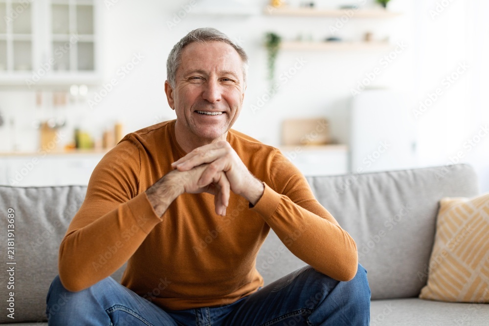 Fototapeta premium Smiling Elderly Caucasian Man Sitting Comfortably On Couch At Home, Displaying Joyful Emotion. Enjoying A Relaxing Moment In A Cozy Living Room Space.
