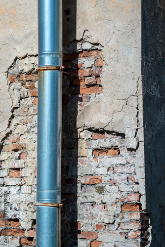 Foto Galvanized metal drainpipe runs down a deteriorating exterior wall where cracked plaster reveals old red bricks, rusted joints and weathered masonry in bright sunlight, suggesting repair needs