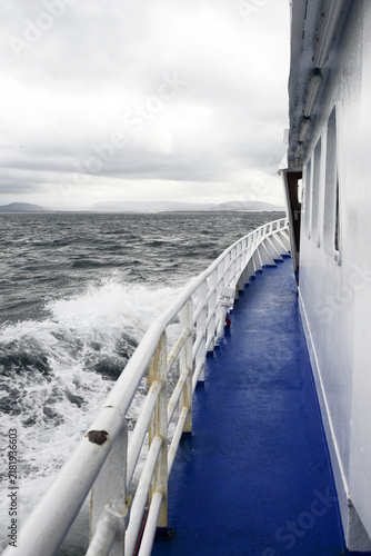 Фотография White railings and a blue deck stretch along the side of a moving ferry churning up sea spray