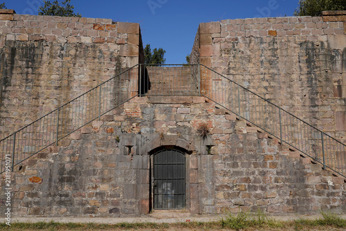 Old stone wall gate with metal bars and symmetrical staircase under clear blue sky in Barcelona