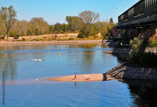 Fotografie Mainz, Rhineland Palatinate, Germany – Rhine river landscape with railway bridge, sandbar and recreational activity on sunny summer day