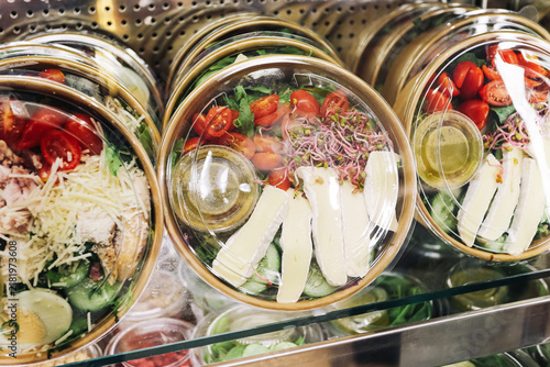 Prepared salads in clear bowls contain tomatoes greens sprouts cheese and dressing under plastic lids. Fresh takeaway meal background with vegetables ready portions and convenient cold food.