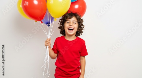Joyful Child with Balloons: A happy young boy with curly hair, dressed in a red t-shirt, beams with excitement while holding a bunch of colorful balloons.