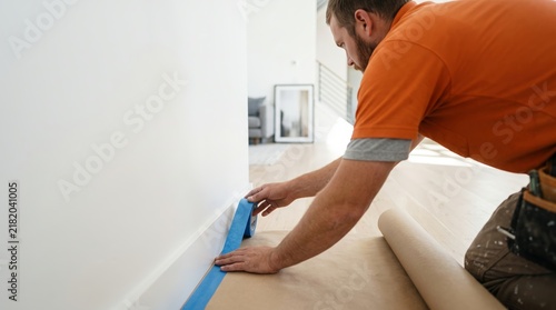 Worker meticulously preparing room for painting by taping baseboards and covering floor