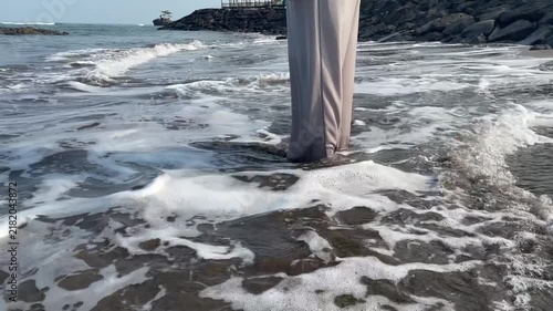 The legs of a woman wearing trousers are splashed by small foamy waves on the beach.