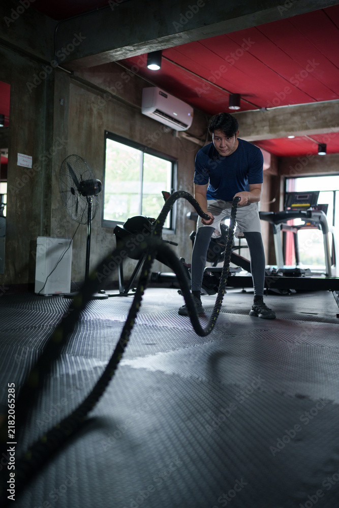 Obraz premium Young Asian man performing intense battle rope exercise in modern fitness center gym interior