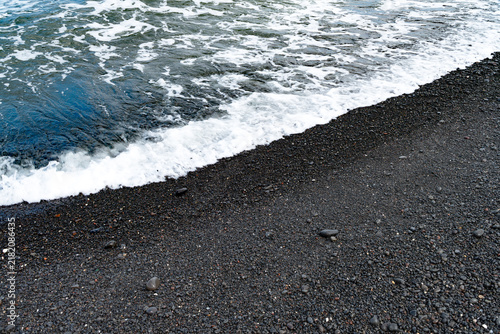 Fotografie Gentle white ocean wave foam washing over dark volcanic sand beach, natural coastal tide line background