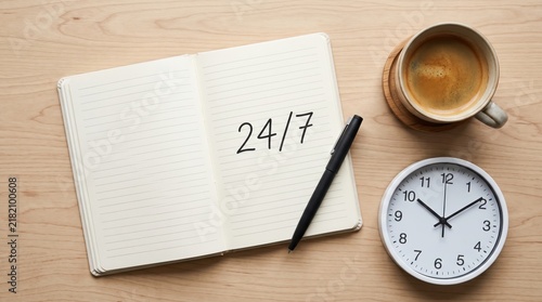 Open notebook with handwritten 24/7 text on wooden desk, coffee cup and clock nearby, round the clock service, business availability, time management, productivity planning