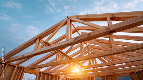 Low angle view of newly constructed wooden roof truss system against clear blue sky with lens flare from bright sunshine.